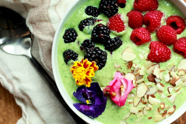 a vibrant green smoothie bowl topped with raspberries, blackberries, almonds and edible flowers on top of a wooden board with fresh berries and a spoon on the side.