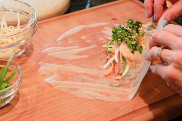 Hands wrapping a fresh spring roll with shrimp and veggies on top of a wooden board.
