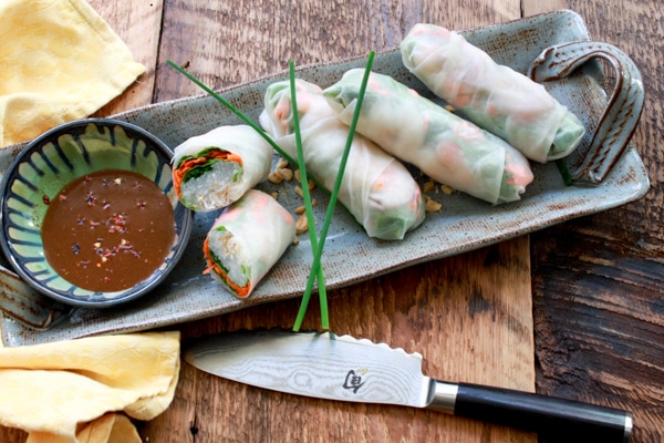 Vietnamese shrimp spring rolls on a long gray platter with a small bowl of peanut sauce on the side on top of a wooden board with a knife and napkin.