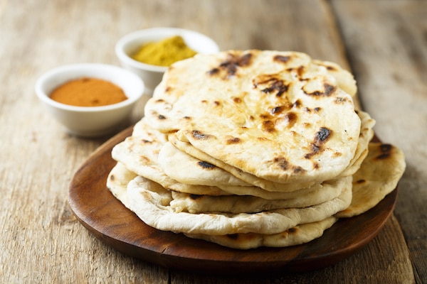 Indian naan bread stacked on a wooden board with spiced in the background
