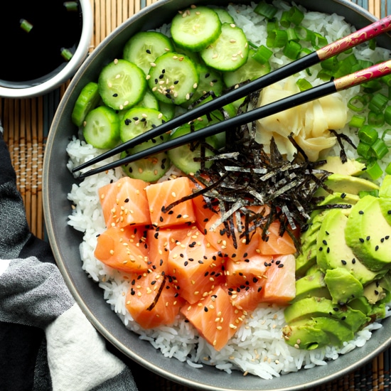 Salmon sushi bowl featuring chunks of sushi-grade salmon, sushi rice, sliced avocados, cucumbers, and seaweed strips with chopsticks.