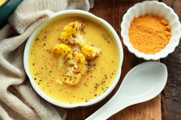vibrant cauliflower soup in a white bowl with a white spoon and turmeric powder on the side