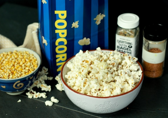 A white bowl of popcorn on a black board with two seasonings behind and a bowl of kernels on the side.