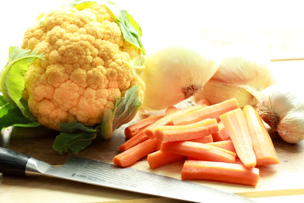 a head of orange cauliflower, carrot sticks, onions, and garlic on a wooden board with a chefs knife