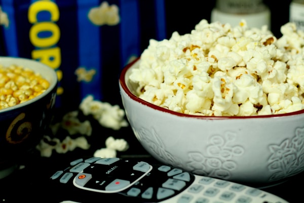 A white bowl with popcorn inside with a TV remote control in front and a bowl of unpopped kernels on the side.