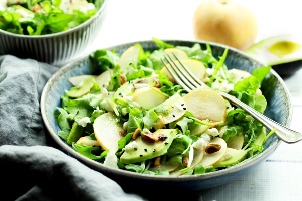 Two bowls of arugula salad topped with Asian pears and avocados with dressing on the side