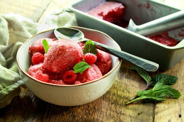 a white bowl of raspberry sorbet and spoon in a white bowl on a wooden board with a metal pan filled with sorbet