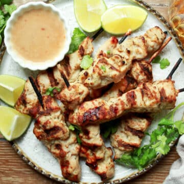 Tender satay chicken pieces cooked on wooden skewers on a white plate with a side of peanut sauce and fresh lime wedges