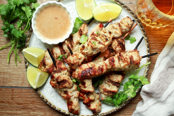 Tender satay chicken pieces cooked on wooden skewers on a white plate with a side of peanut sauce and fresh lime wedges