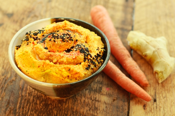 a bowl of roasted carrot and ginger hummus on a wooden board with raw carrots and fresh ginger on the side