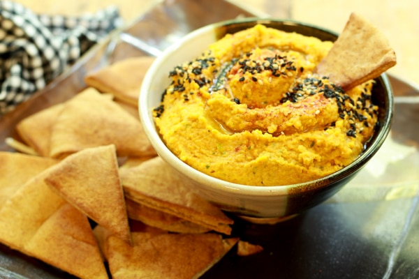 a small bowl of roasted carrot and ginger hummus with a pita chip inserted and pita chips on the side