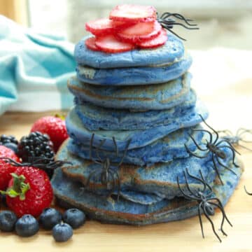 a stack of blue pancakes on a wooden board with vibrant berries on the side and plastic spiders crawling up the stack