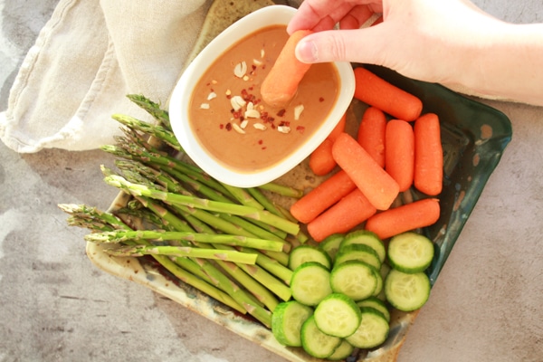 a woman dipping a carrot into a small white bowl of peanut sauce that is placed on a tray with fresh vegetables