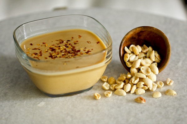 Spicy peanut dressing inside a clear glass bowl with raw peanuts on a gray board.