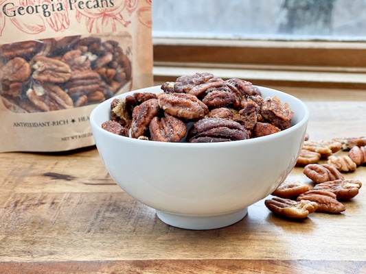 Spiced whole pecans in a white bowl with a bag of pecans and loose pecans on the side on a wooden board.