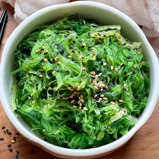 A white bowl filled with Japanese seaweed salad with black chopsticks and a linen napkin on the side on top of a wooden board