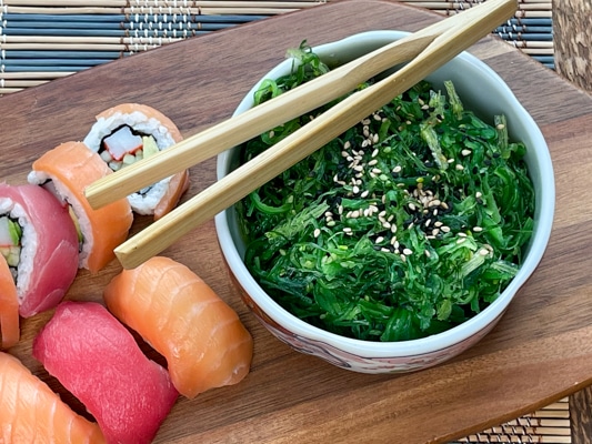 A bowl filled with Japanese seaweed salad with wooden tongs on top and assorted sushi on the side, on a wooden board