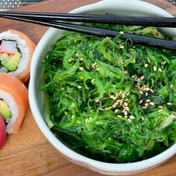 A bowl filled with Japanese seaweed salad with black chopsticks on top and assorted sushi on the side, on a wooden board.