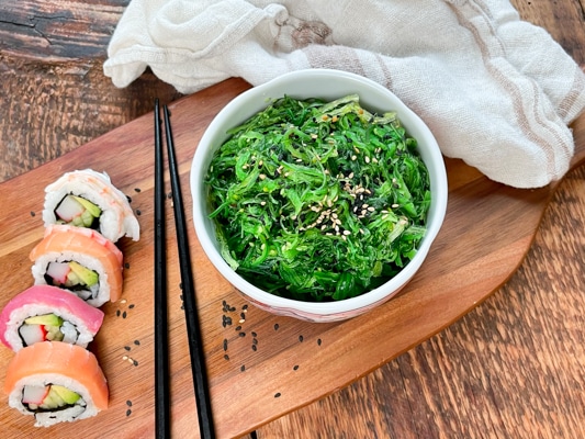 A bowl filled with Japanese seaweed salad with black chopsticks and assorted sushi on the side, on a wooden board with a linen napkin