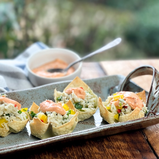 A long gray serving platter with baked wonton cups filled with crab salad and a dollop of colorful aioli on top on a wooden board and a small white bowl of aioli behind it.