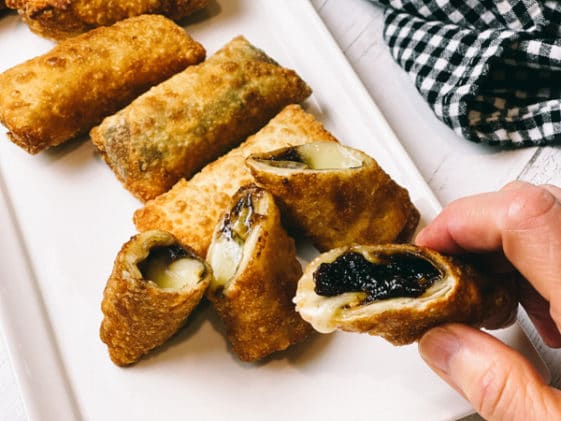 A woman's hand holding a crispy, golden spring roll filled with brie and fig with more spring rolls on a long white serving platter.
