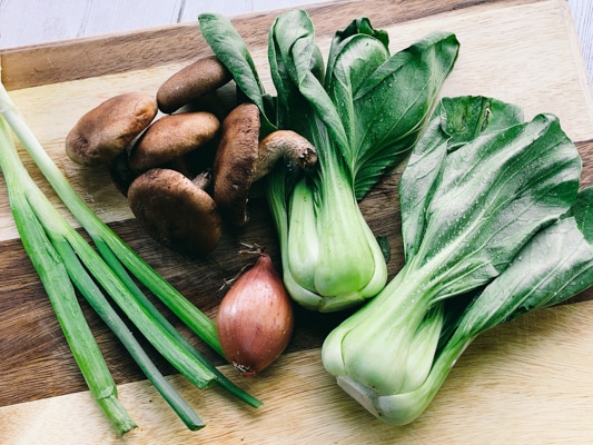 Fresh vegetables on a wooden board including green onions, shiitake mushrooms, shallots, and baby bok choy