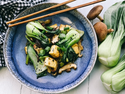 Stir-fry miso shiitake mushrooms, tofu, and baby bok choy on a blue plate with wooden chopsticks, and fresh baby bok choy and shiitake mushrooms on the side.