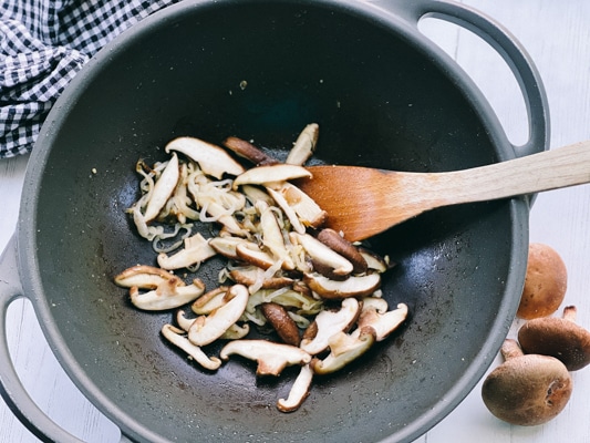 Sliced shiitake mushrooms being stir-fried in a black wok with a wooden spatula inside and raw mushrooms on the side.