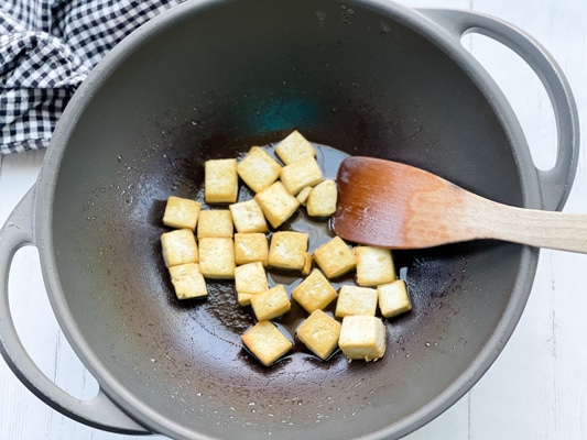 Cubed tofu being stir-fried in a black wok with a wooden spatula on top of a white board
