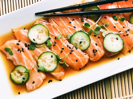 Slices of fresh sashimi salmon on a white platter with sliced cucumbers on top and sprinkles of black sesame seeds, with black chopsticks.