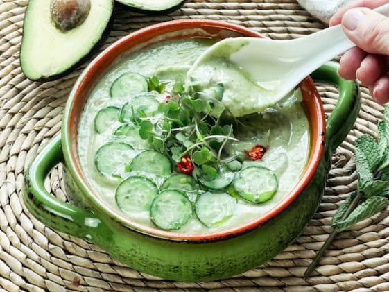 A woman's hand holding a white spoon inside a green bowl filled with cucumber avocado soup, with sliced avocados on the side.