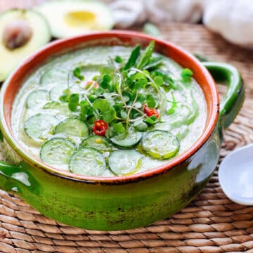 A bowl filled with spicy cucumber avocado soup topped with sliced cucumbers and herbs with a sliced avocado and a white spoon on the side.