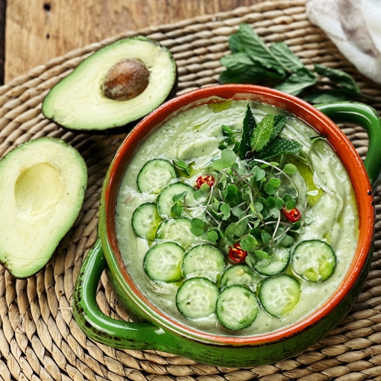 A bowl filled with spicy cucumber avocado soup topped with sliced cucumbers and herbs with a sliced avocado on the side.