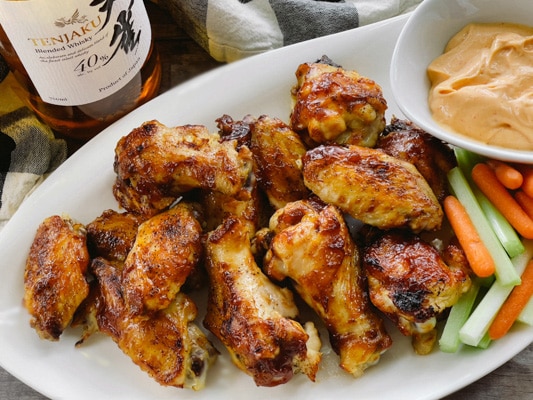 A bottle of Japanese whiskey placed behind a white plate of saucy chicken wings, celery and carrot sticks, and a bowl of aioli dip.