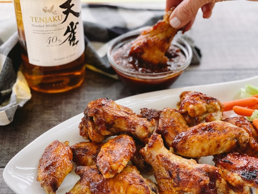 A woman's hand dipping a chicken wing drum into sauce next to a bottle of Japanese whiskey, and a white plate of chicken wings in front.