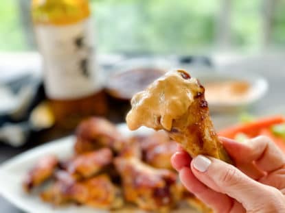 A woman's hand holding a saucy chicken wing drum that's been dipped in an aioli sauce.