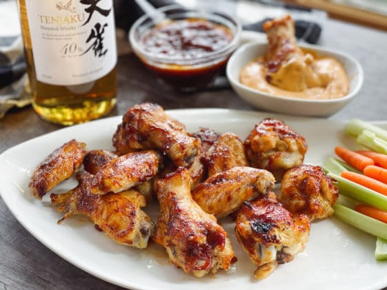 A bottle of Japanese whiskey placed behind a white plate of saucy chicken wings, celery and carrot sticks, with a bowl of aioli dip and extra sauce behind.
