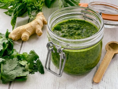 A clear glass bowl of cilantro, mint, basil sauce with a small wooden spoon on the side, and bunch of fresh cilantro and basil leaves, and ginger root, all on top of a white wooden board.