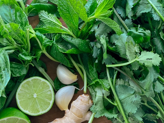 Fresh basil, mint and cilantro leaves, and two lemon halves, ginger root, and garlic cloves on a wooden board.