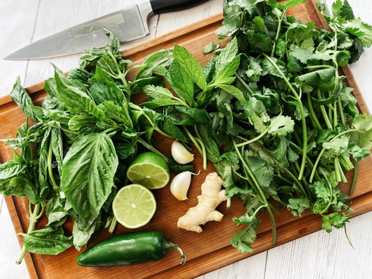 Fresh basil, cilantro, mint, and cilantro leaves on top of a wooden cutting board, along with a jalapeno pepper, fresh lime halves, and garlic cloves.