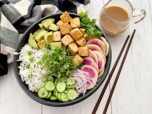 A black bowl filled with miso tofu poke bowl ingredients and white rice, with chopsticks and a clear jar of miso ginger dressing on the side.
