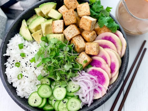 A black bowl filled with miso tofu poke bowl ingredients and white rice, with chopsticks and a clear jar of miso ginger dressing on the side.