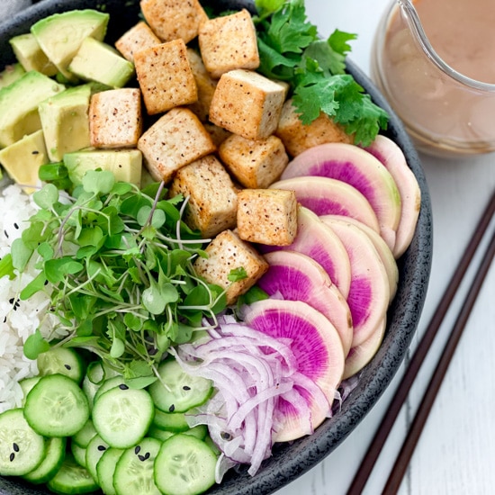 A black bowl filled with miso tofu poke bowl ingredients and white rice, with chopsticks and clear jar of miso ginger dressing on the side.