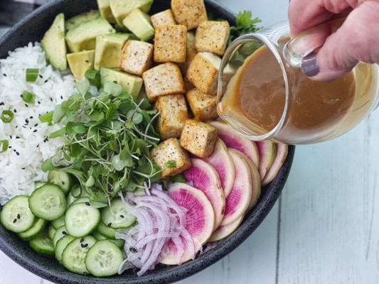 A miso tofu poke bowl in a black vessel with a woman pouring miso ginger dressing on top.