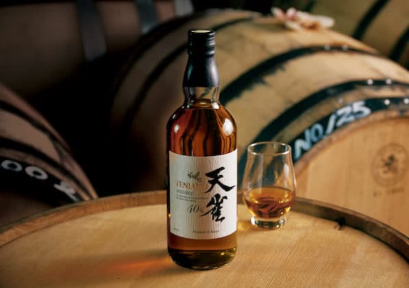 A bottle of Tenjaku whiskey from Japan on top of a wooden whiskey barrel with other barrels behind it.