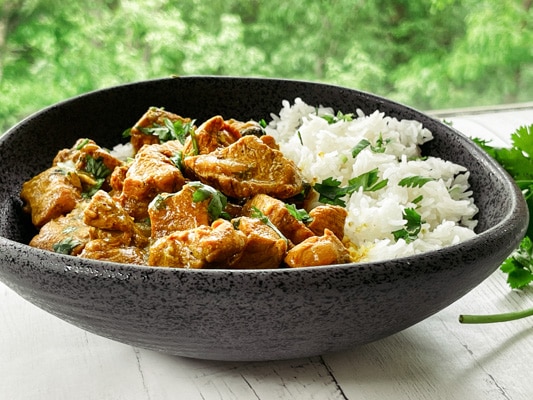A black bowl of Thai curry chicken with white rice and fresh cilantro on the side, on top of a white board.