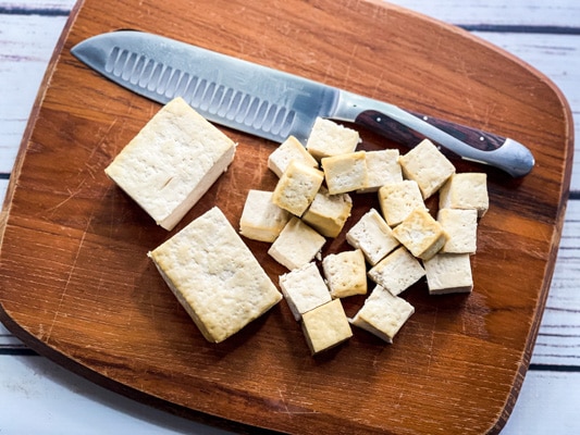 Cubed extra firm tofu on a wooden cutting board with a chefs knife along side.