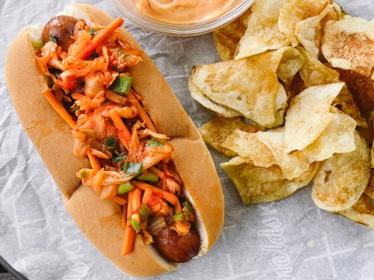 A hot dog in a bun topped with kimchi slaw with potato chips and aioli on the side.