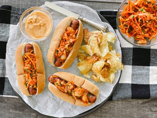 Three hot dogs in buns topped with kimchi slaw with potato chips, extra slaw and aioli on the side on top of a checkered napkin.