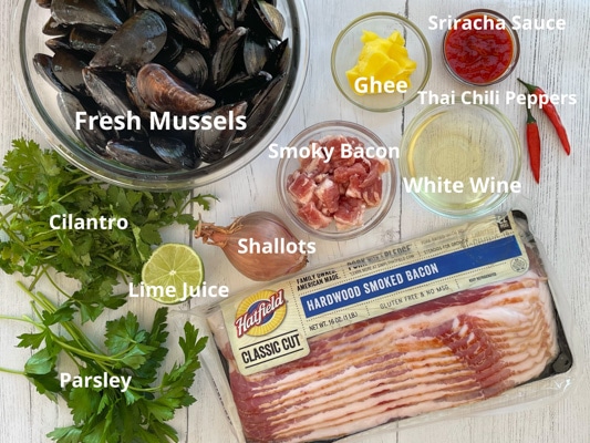 Ingredients for steamed mussels on a white board including a package of Hatfield hardwood smoked bacon, fresh green herbs, shallots, fresh mussels in a bowl, cut bacon, white wine, ghee, Thai chili peppers, and sriracha sauce.